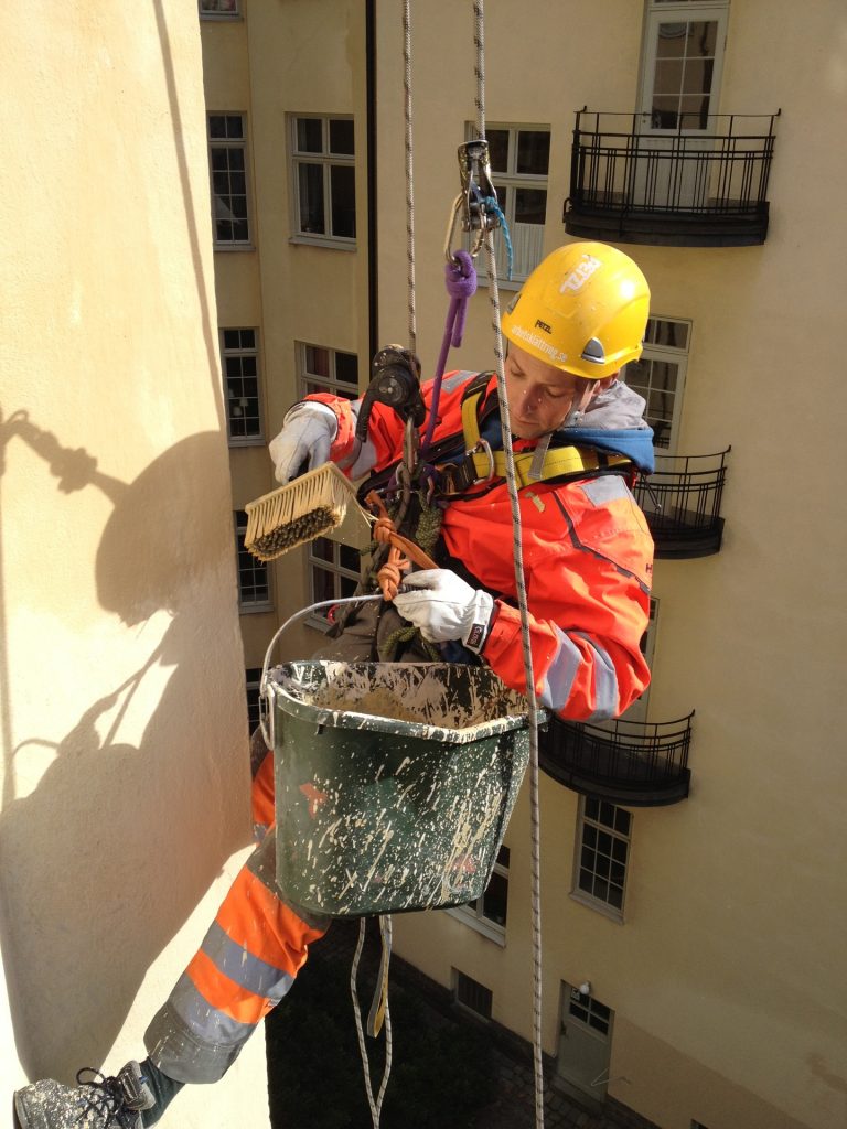 Medarbetare i Stockholm som är upphissad med lina i varselkläder målar husfasad